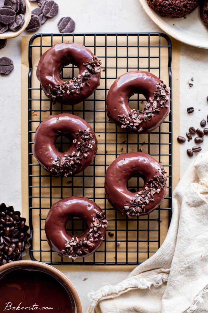 Five chocolate-dipped chocolate cake donuts on a black cooling rack. Each donuts topped with cacao nibs and crushed coffee beans, with a white napkin on the right side. All on a white background.