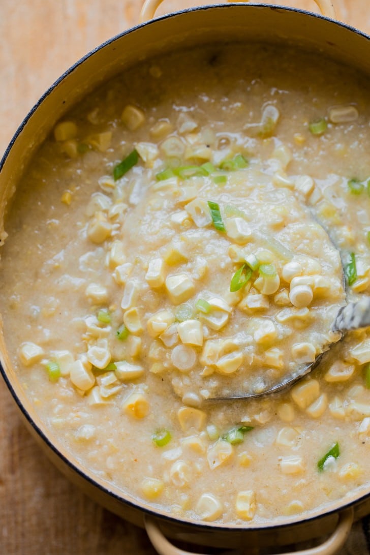 A pot full of vegan corn chowder being ladled out, garnished with fresh sweet corn kernels and green onions.