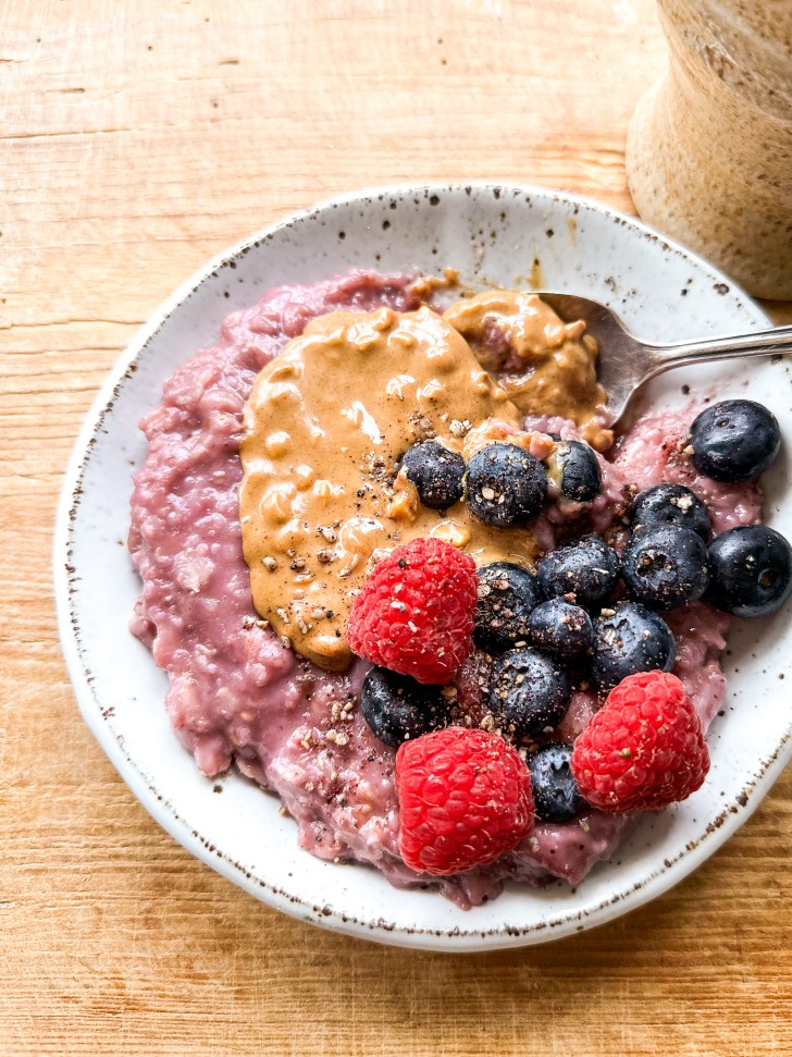 White ceramic bowl filled with purple berry oatmeal and topped with peanut butter, blueberries, raspberries, and cinnamon.